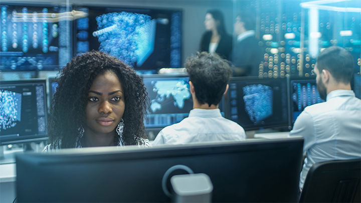 a woman with a dark skin tone sits in front of a computer in a busy SOC (security operations center)